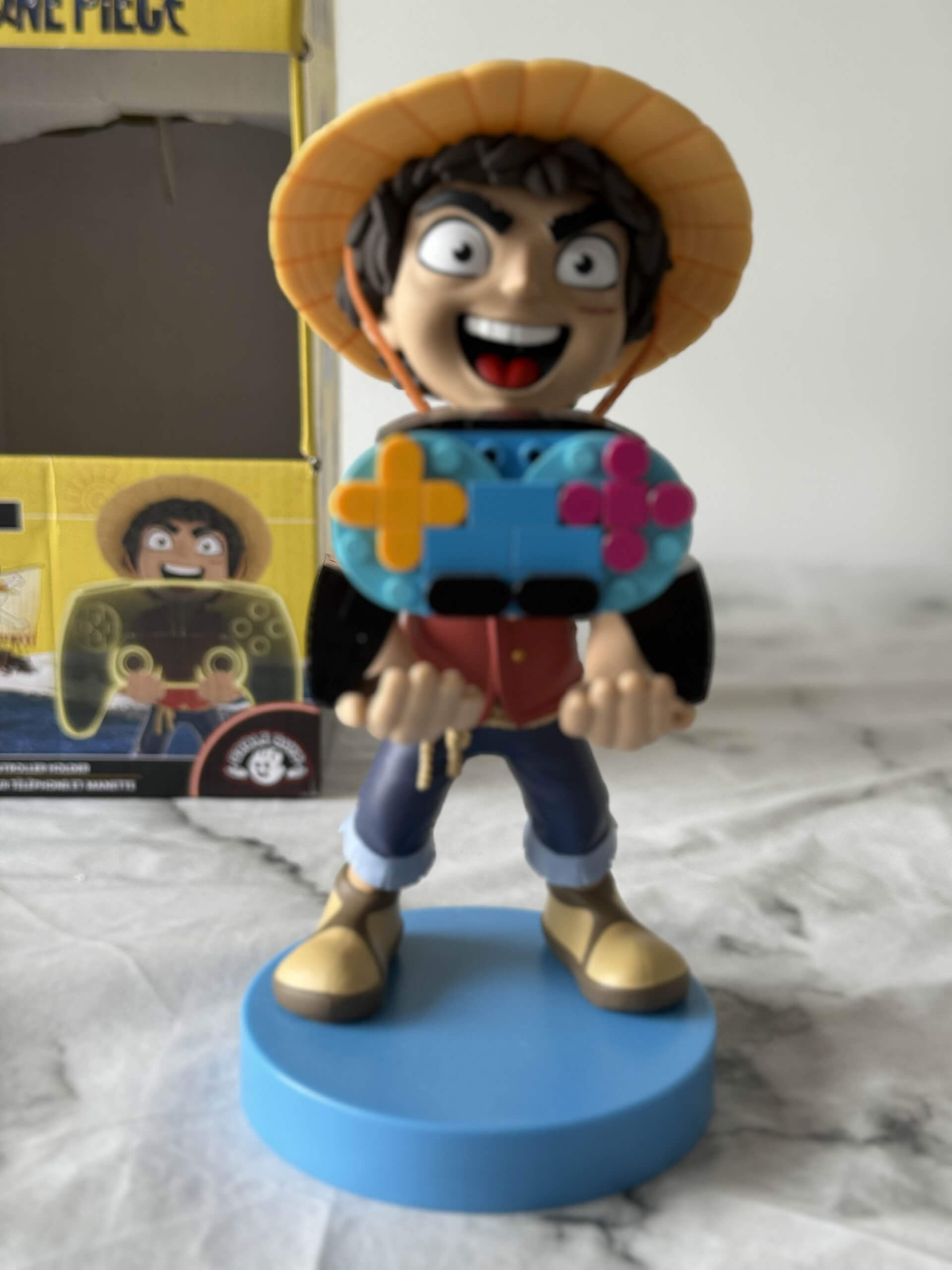 A collectible figurine of a character wearing a straw hat, holding a colorful gaming controller, placed on a blue base with a box in the background.
