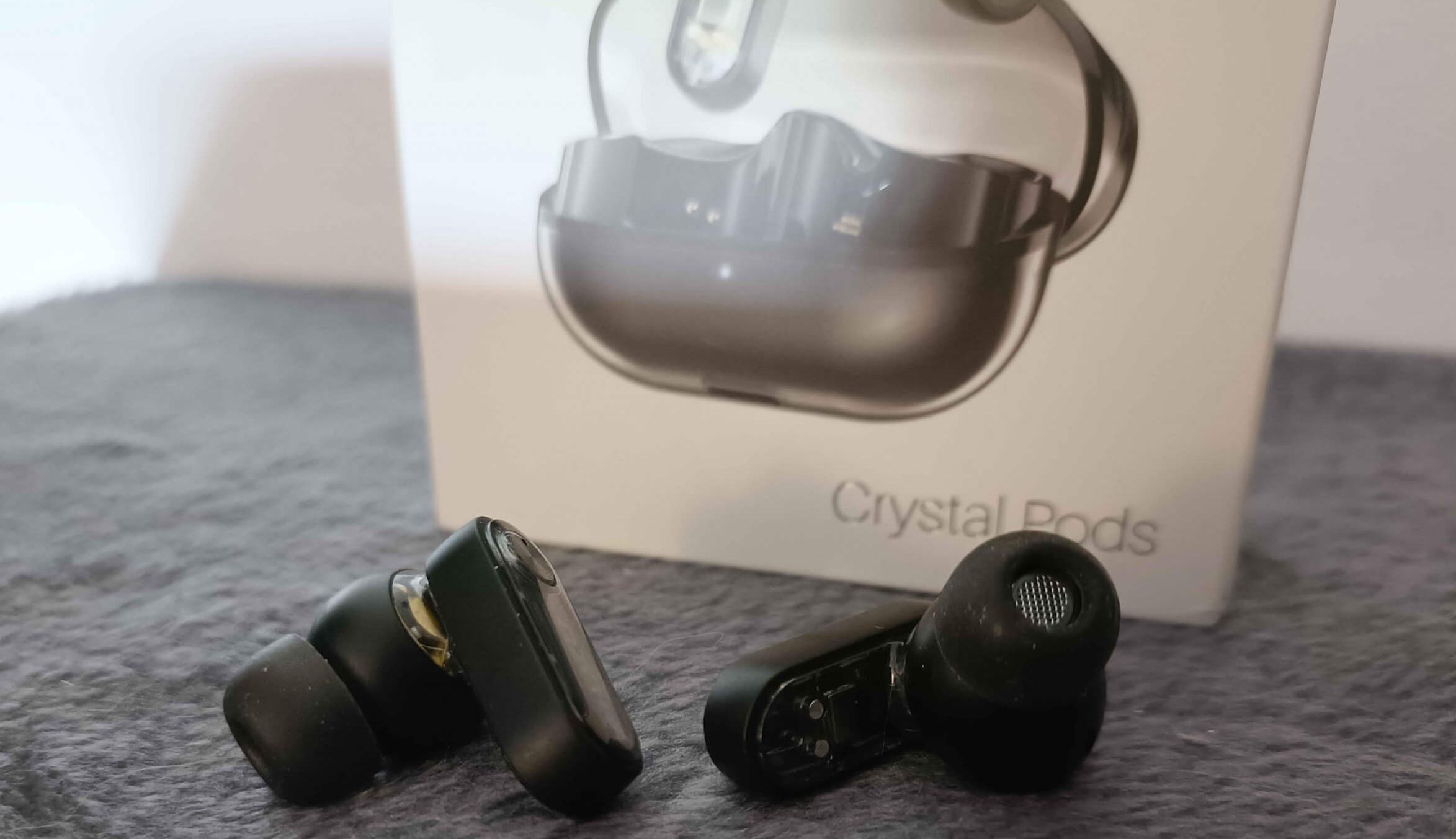 Two black wireless earbuds are displayed beside a box labelled "Crystal Pods" on a textured grey surface.