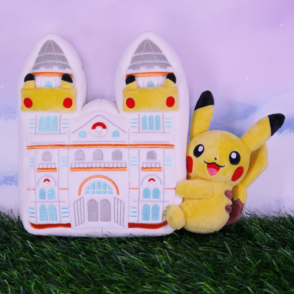 A plush Pikachu sits beside a soft castle-shaped cushion, decorated with Pokémon motifs, on a grassy surface against a pastel background.