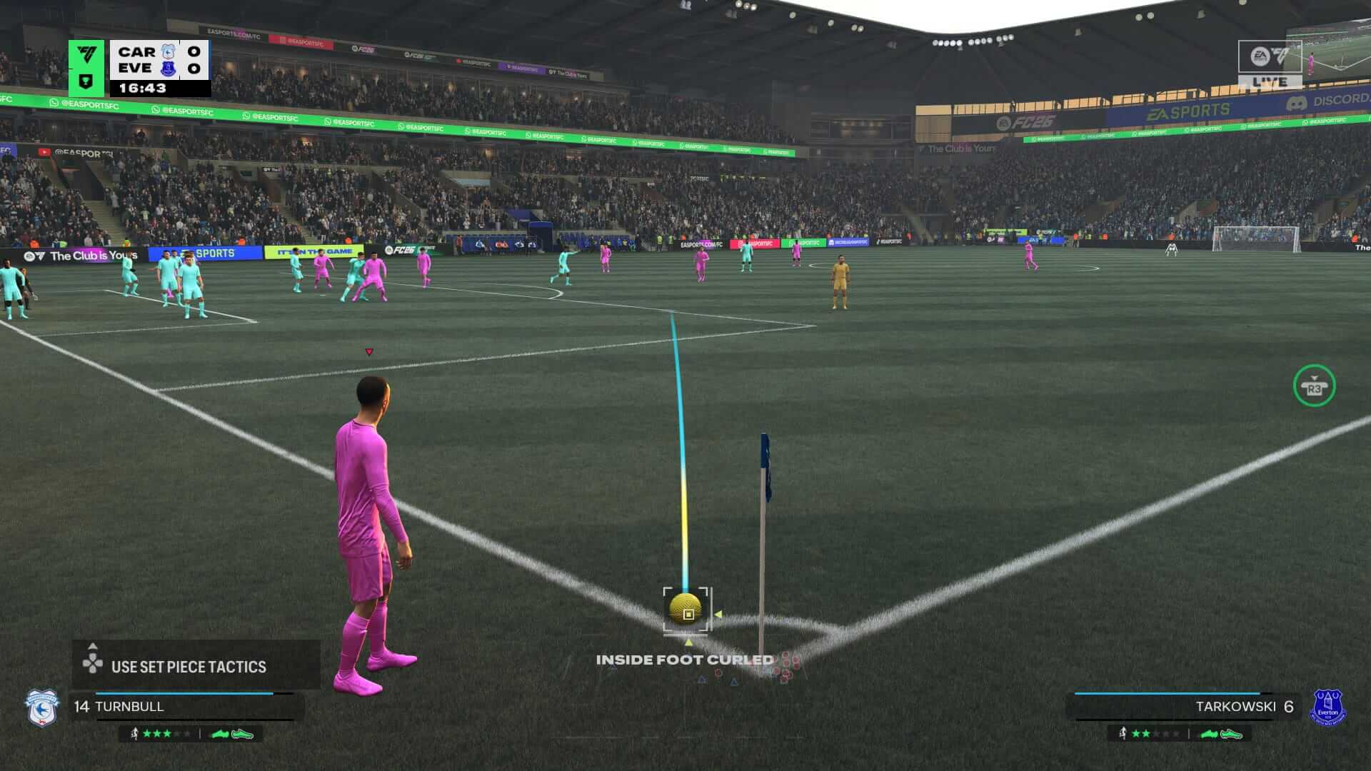Screenshot from EA Sports FC 26. A corner routine shows a player in pink preparing to kick. The field is full of players in teal and pink, and the referee in Orange. The stadium is packed with fans.