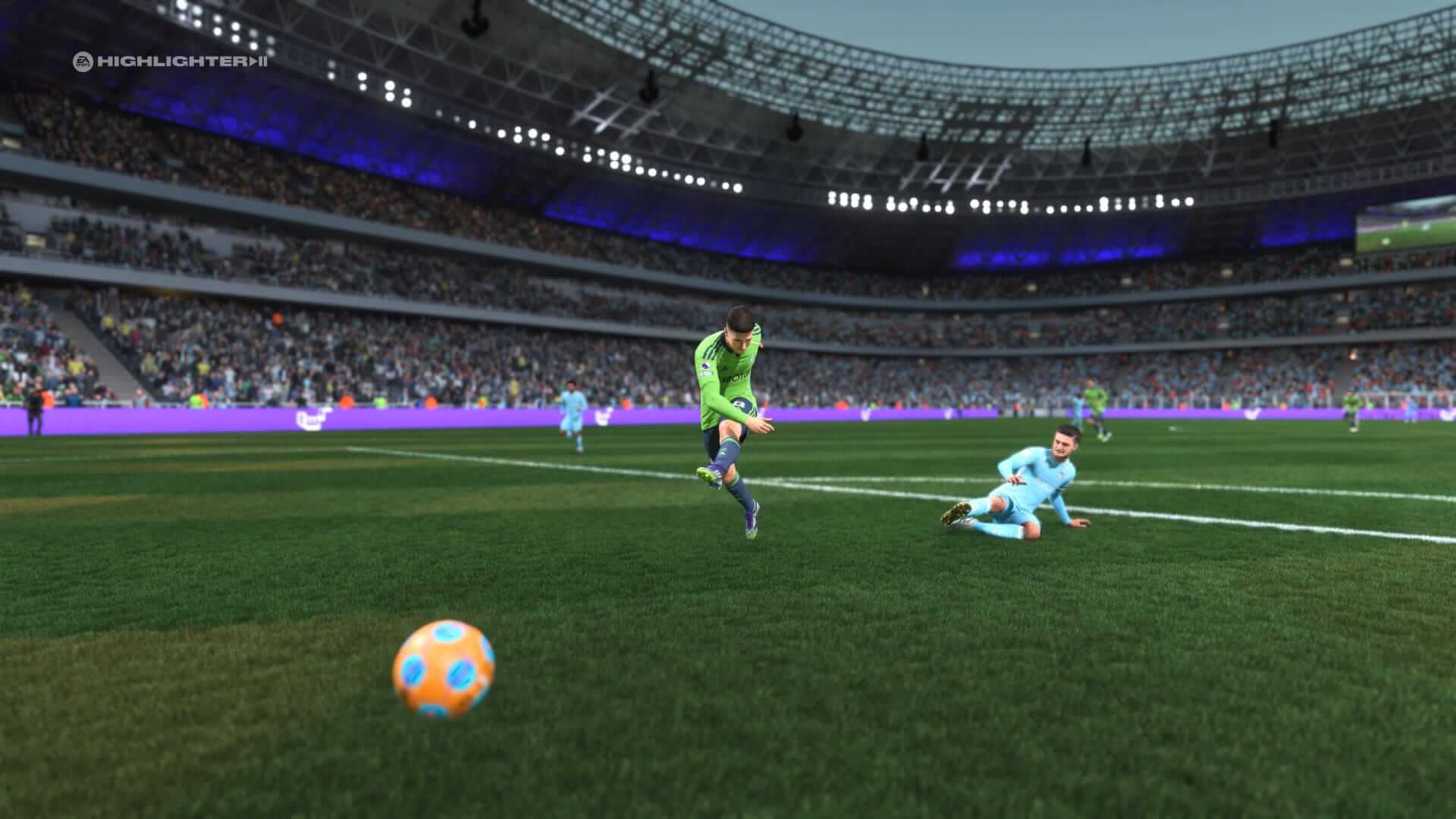 Screenshot from EA Sports FC 26. A football player in a lime green kicks a blue and orange ball towards the goal as an opponent in light blue slides on the grass. The viewpoint is from the goalkeeper's perspective.