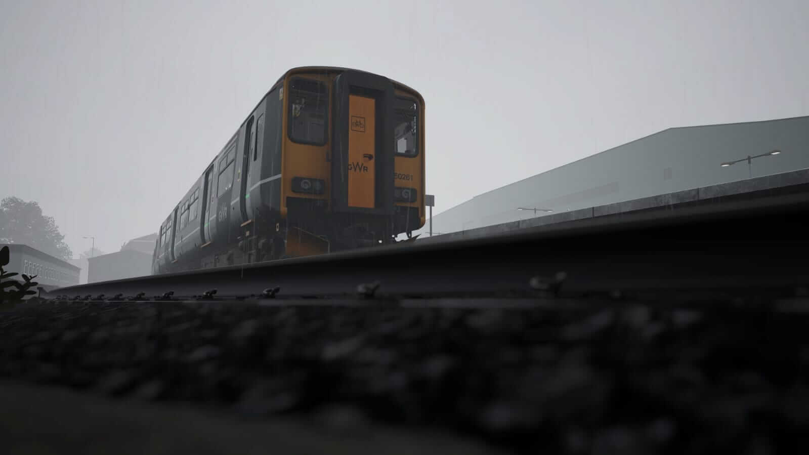 Screenshot from Train Sim World 6. A low-angle view of a train on a misty day, with overcast skies. The yellow front contrasts with the grey surroundings, evoking a quiet, sombre mood.
