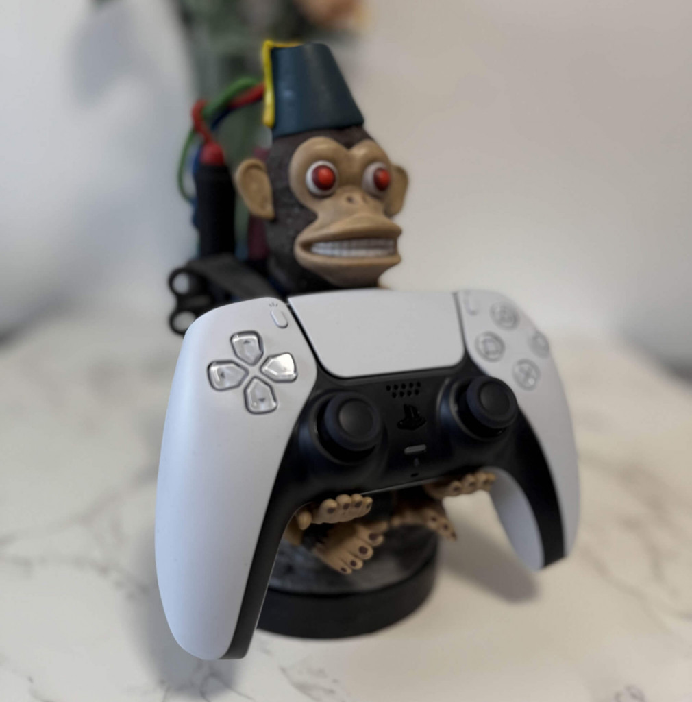 A white gaming controller sits in the hands of a quirky monkey bomb cable guys figurine. In the background is a vase of flowers.