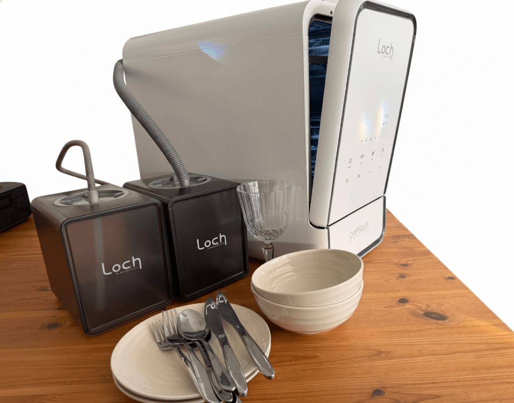 A modern white water dishwasher with two black containers, a glass, and ceramic bowls and utensils on a wooden table.