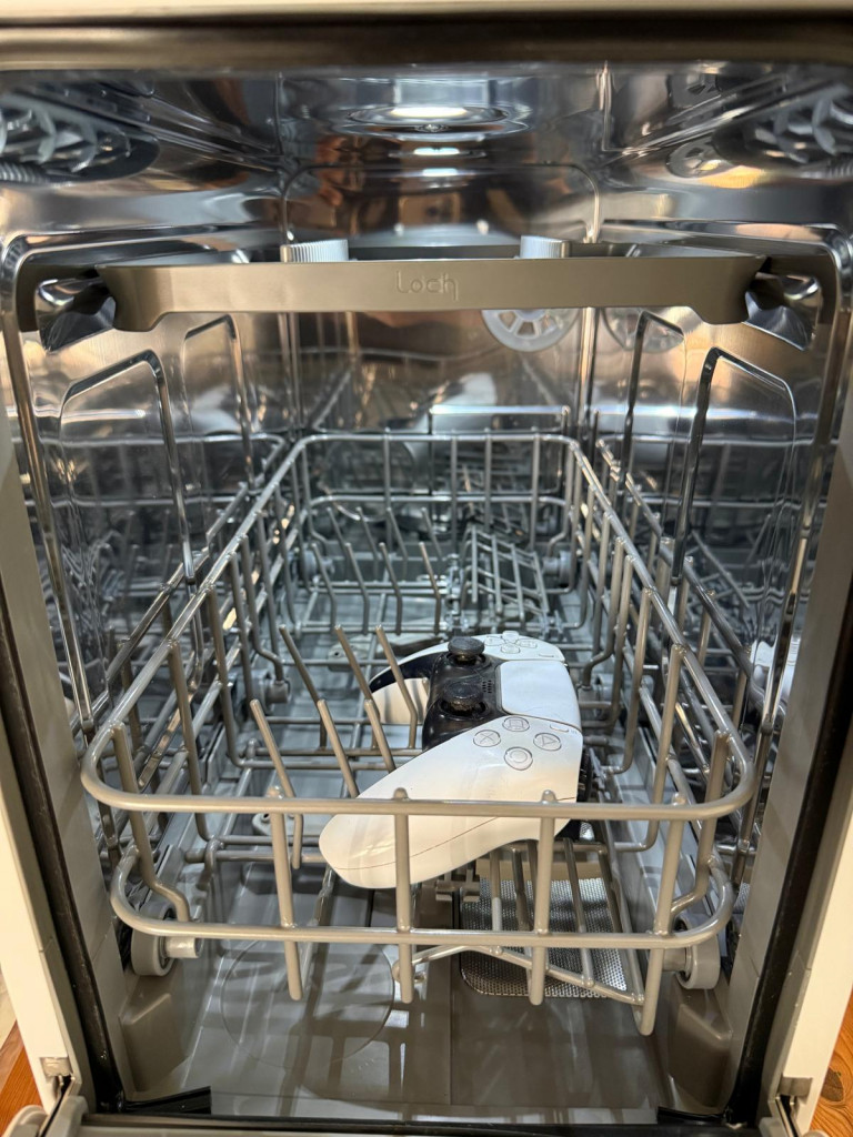 A white gaming controller sits inside an empty dishwasher's wire basket, highlighting an unconventional cleaning choice.