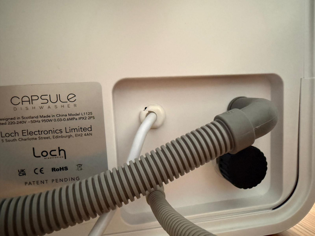 Close-up of the Capsule dishwasher's side panel showing model information, and connected hoses and fittings.