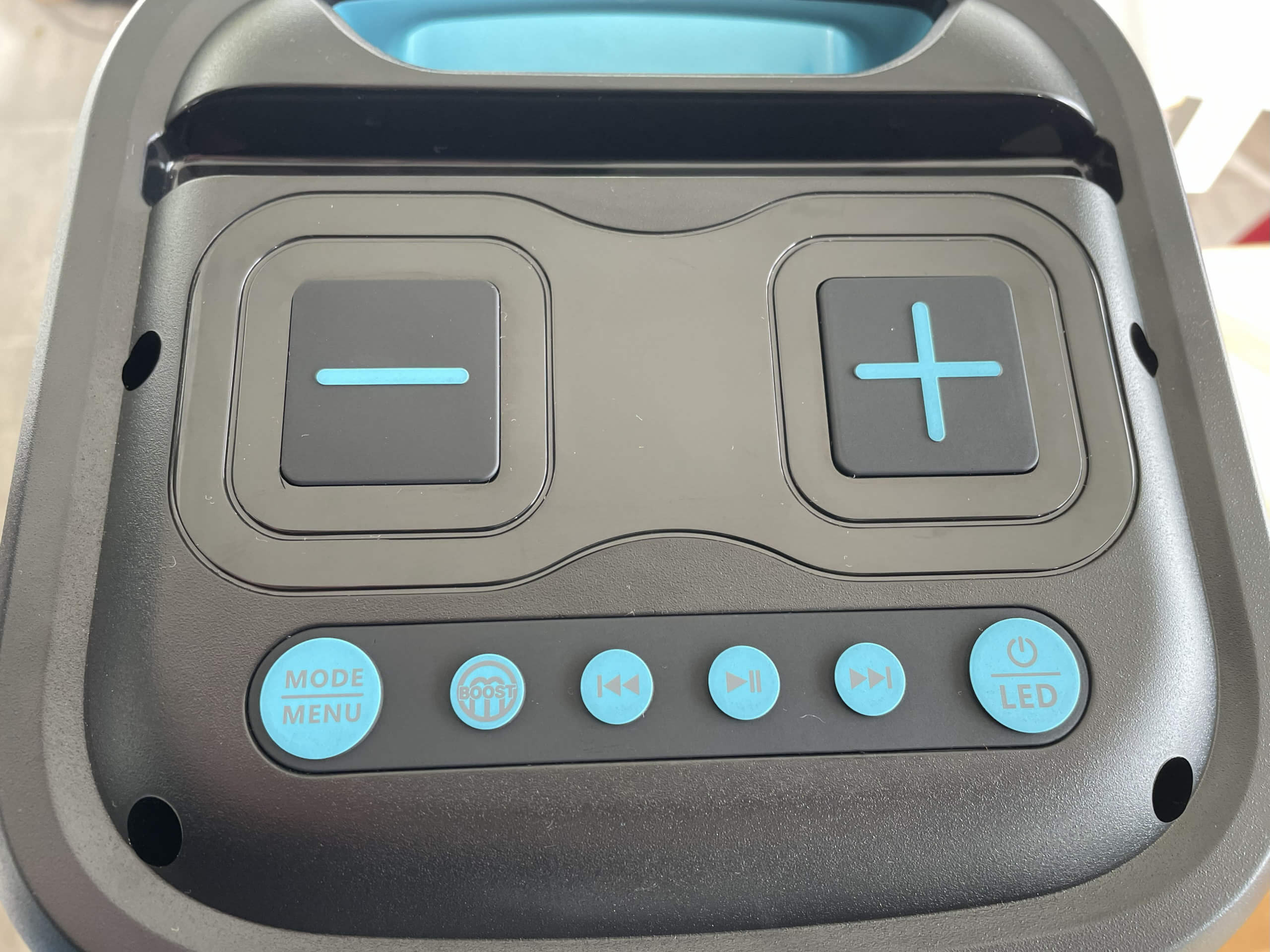 The Pulse 1 control panel, viewed from above. 2 large, square buttons, 1 with a minus symbol and 1 with a plus, situated above a row of smaller, circular buttons. Circular buttons from left to right are mode/menu, boost, back, plau/pause, forward, and power/LED. The speaker has a black and light blue colour scheme.