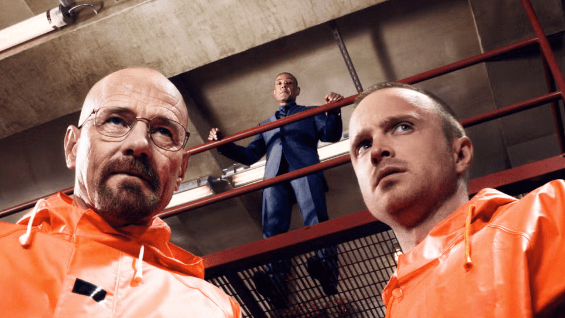 Two men in orange protective suits look serious, standing below a man in a suit on a walkway. The setting is industrial, with tense, dramatic lighting.