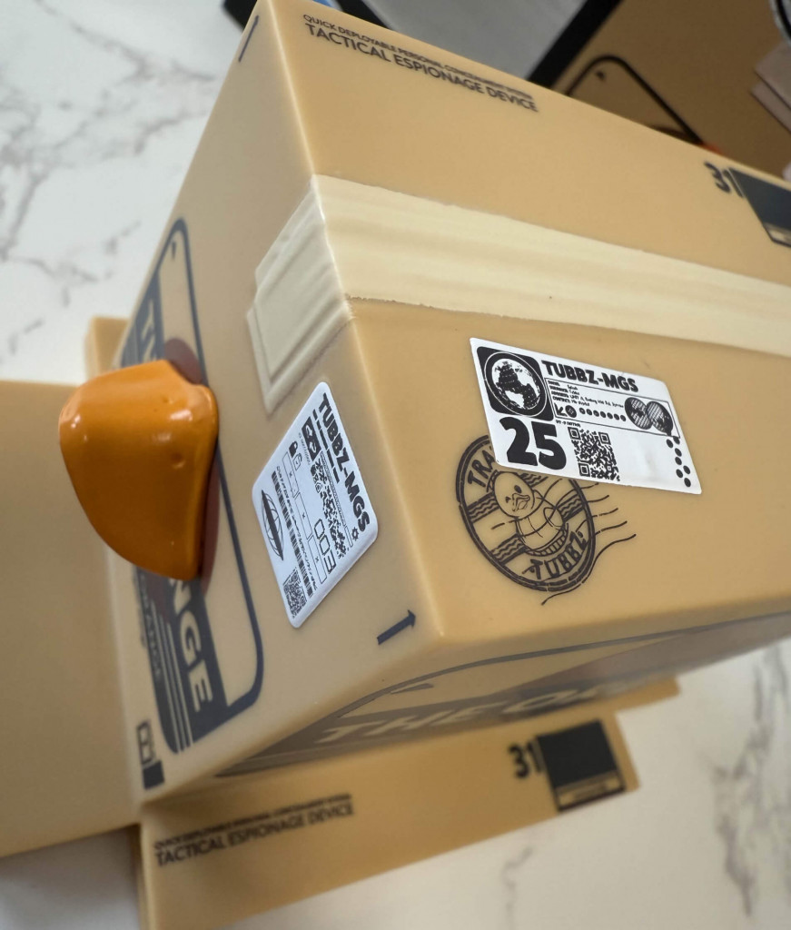 A close-up of the Orange Box MGS Tubbz figure. A tan box labelled "Tactical Espionage Device," featuring an orange duck bill sticking out and various stickers on its surface.