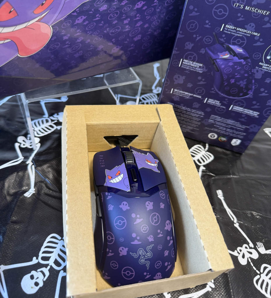A purple Razer gaming mouse featuring playful Gengar designs, displayed in its cardboard packaging on a skeleton-patterned surface.