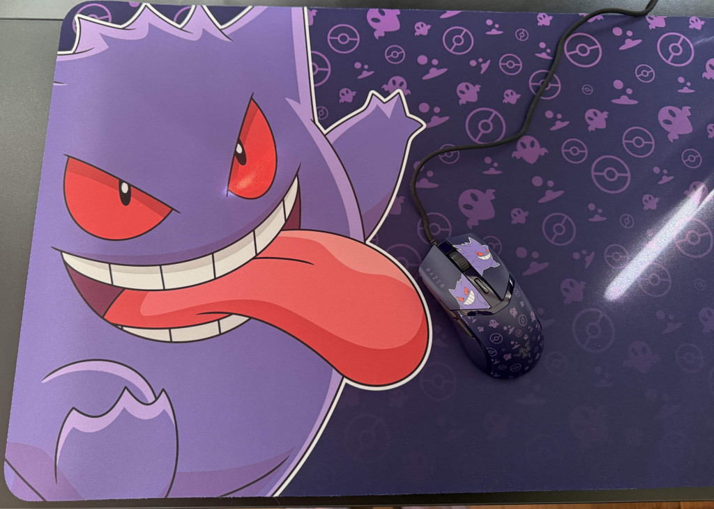 A purple razer cobra gaming mouse with Gengar graphics and playful designs rests on a matching mouse pad featuring a spooky aesthetic. The Gengar on the mouse mat appears to be licking he mouse.