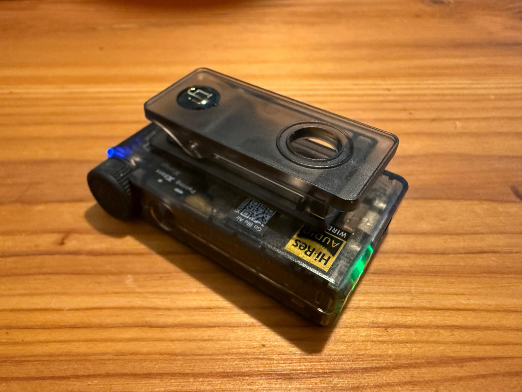 Translucent audio DAC featuring a chrono dial button, and a magnetic belt clip rests on a wooden surface. Blue and green lights are visible.