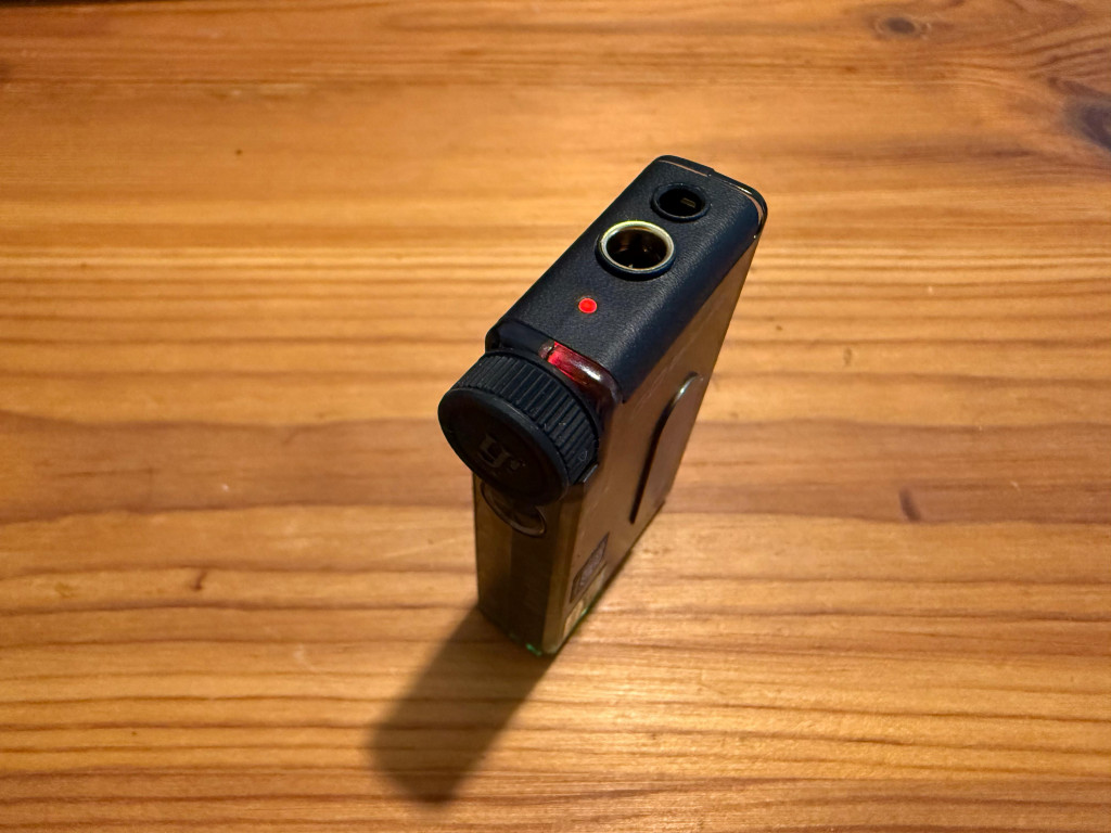 A compact audio device with a black dial and input ports on top rests on a wooden surface. It has a small red indicator light, suggesting it's powered on.