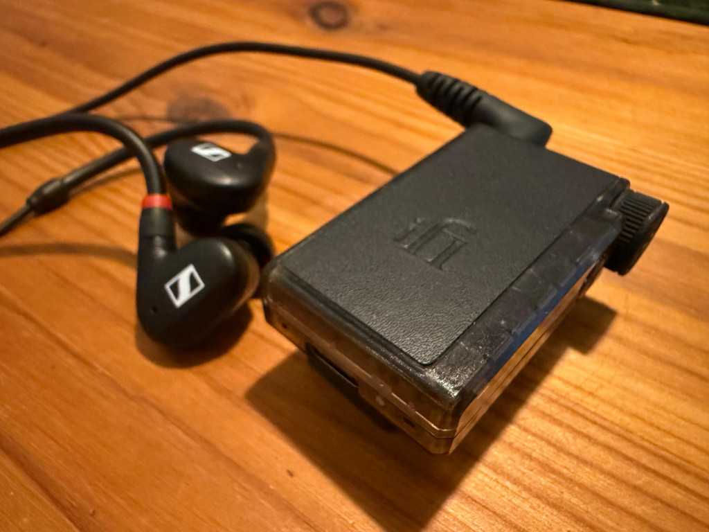 A pair of black in-ear headphones with a red accent rests beside a compact black audio DAC device on a wooden table, conveying a tech-savvy mood.