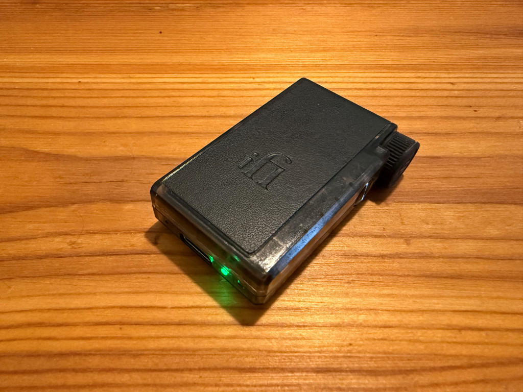 A small black audio device with the iFi logo rests on a wooden table. It has a visible green indicator light, creating a modern and sleek appearance.