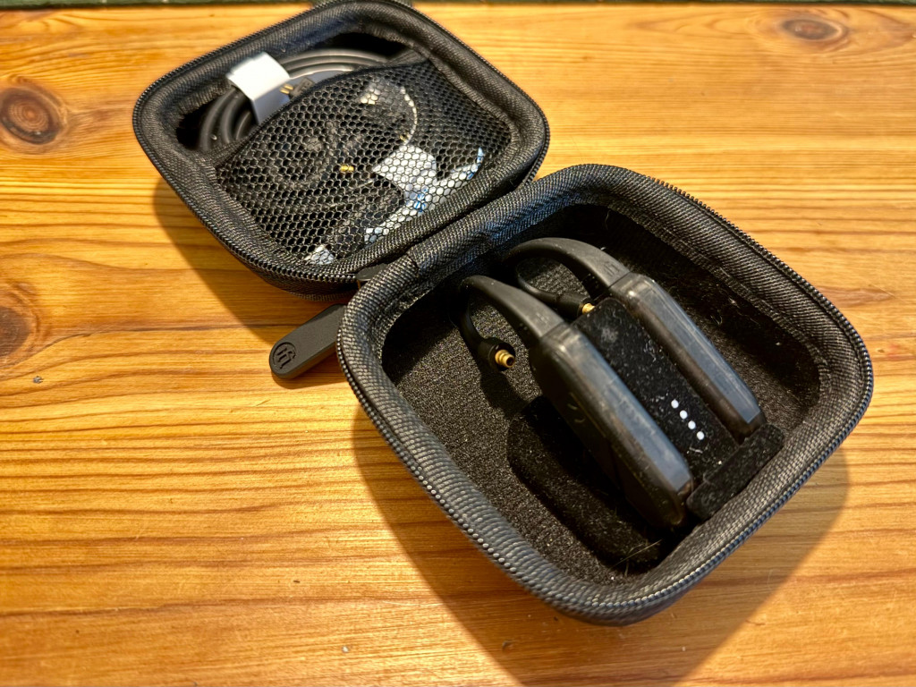 A small black zippered case is open on a wooden surface, displaying two in-ear monitors with attached cables. A mesh pocket contains additional accessories.