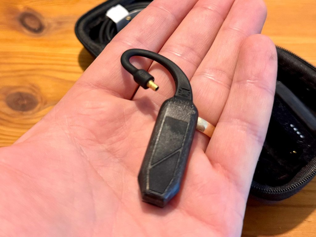 Close-up of a hand holding a small black ifi go pod air device with a curved cable and a gold connector, set against a wooden surface with a black case nearby.