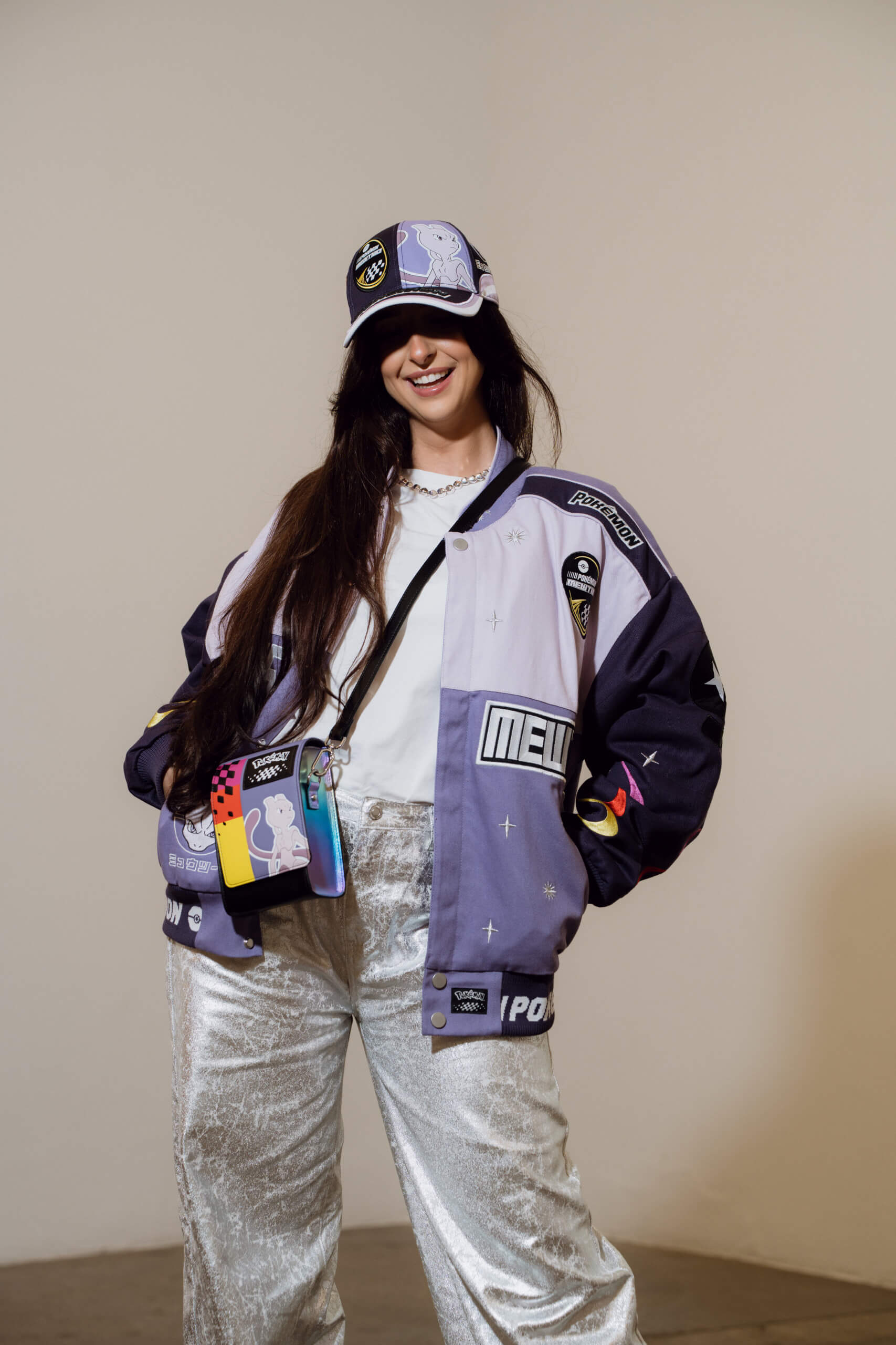 A femme presenting person wears a Mewtwo inspired sports jacket with grey jeans and accessorised with a mewtwo patterned cross body bag from the Pokémon x JimmyPaul by Difuzed line.
