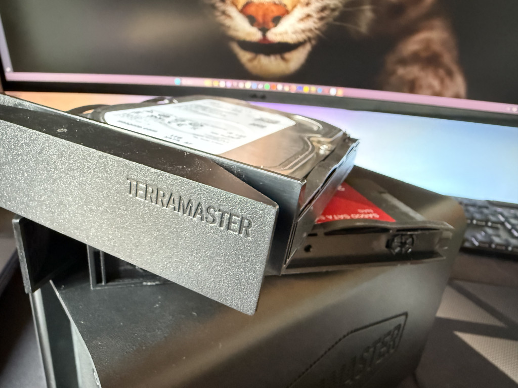 Close-up of a Terramaster hard drive enclosure with two hard drives protruding. A computer screen displays a close-up image of a big cat's face.
