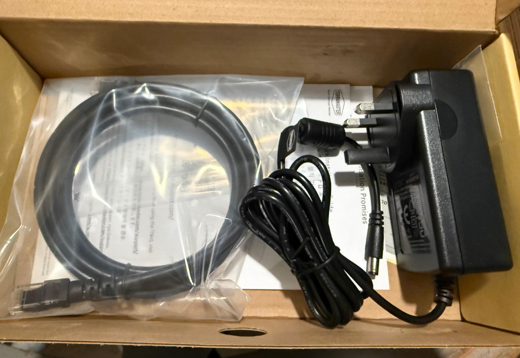 A cardboard box contains a coiled black cable in a plastic bag and a black power adapter with a plug. A paper document is visible underneath.