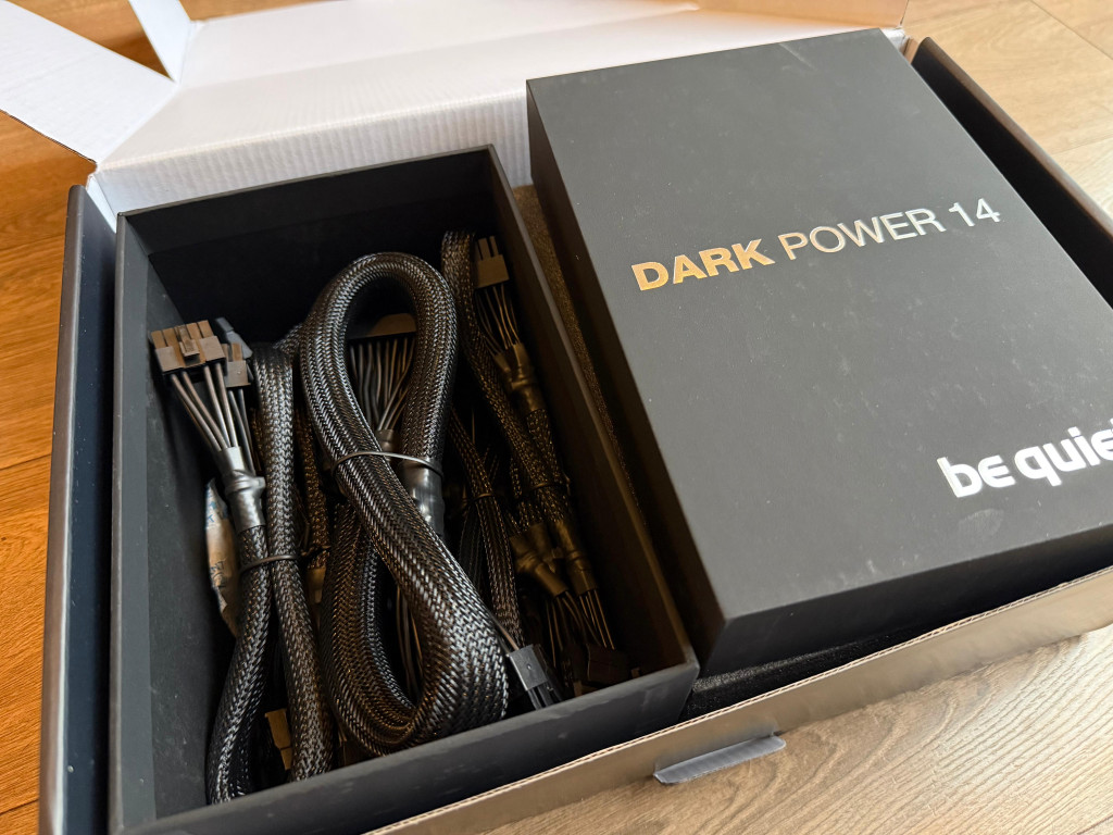 Box of computer cables with a "Dark Power 14" power supply by be quiet! The packaging is open, displaying neatly coiled black cables on a wooden surface.