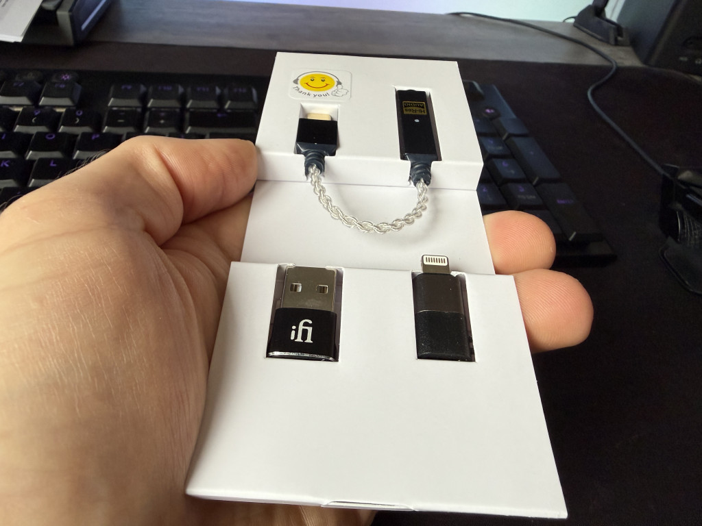 Close-up of a hand holding a white package with various adapters and a small cable inside. A smiley sticker and a keyboard are in the background.