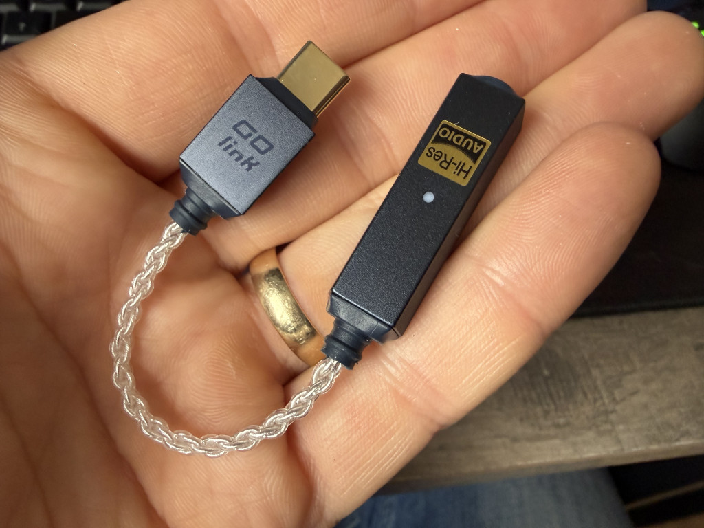 Close-up of a hand holding a small audio dongle with a braided cable. Connector labelled "Go link," other end says "Hi-Res Audio." Warm lighting on a soft background.