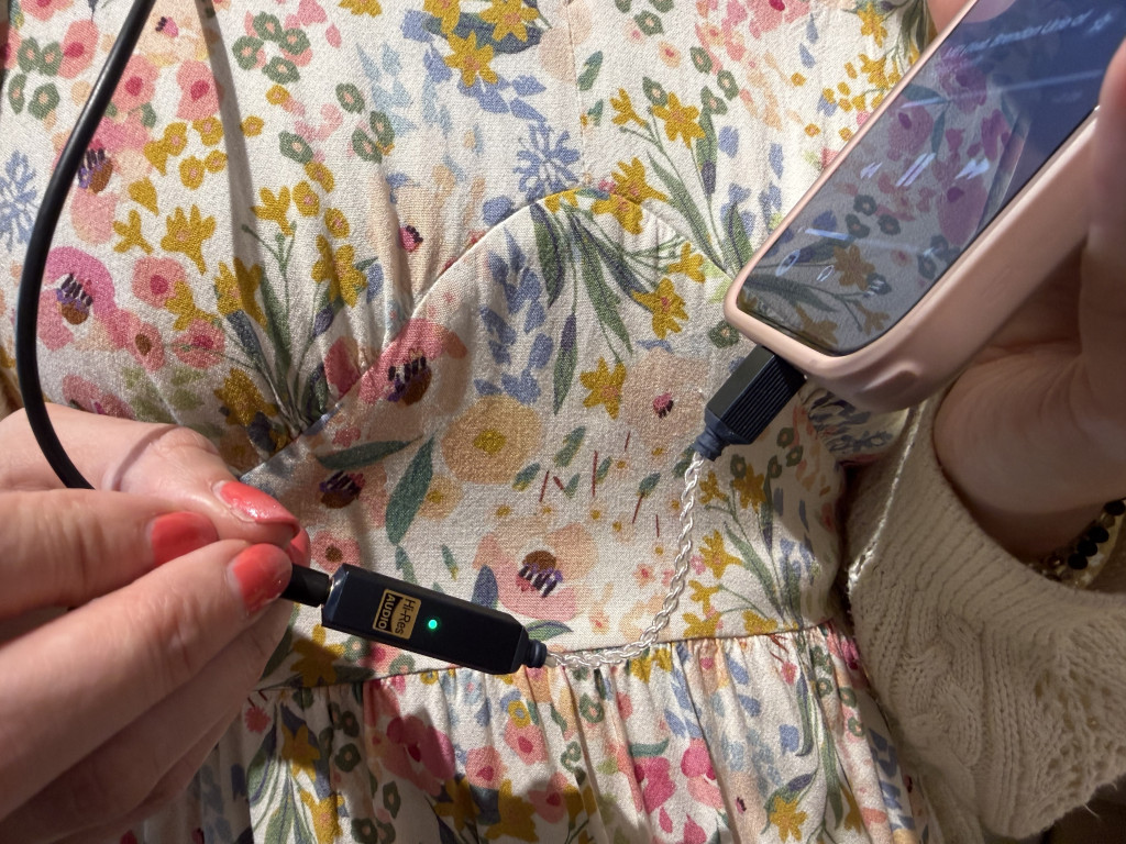 Close-up of a person in a floral dress holding a phone with an iFi GO link 2 plugged in along with headphones. There is a green LED light indicator. Bright, casual setting.