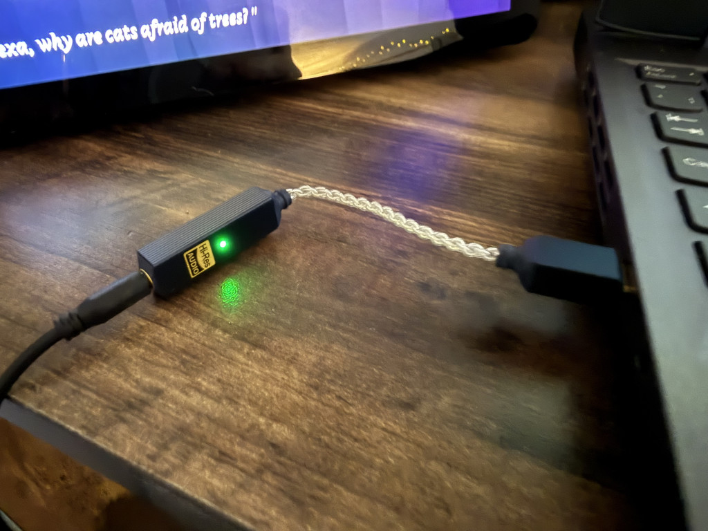 A iFi GO link 2 with a glowing green light is connected to a laptop on a wooden desk. A screen displays text in the background.