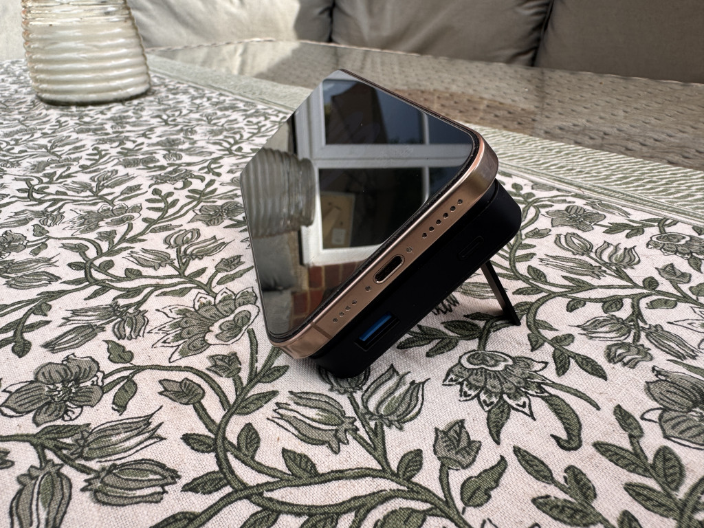 Smartphone resting at an angle on a stand, reflecting a window. It’s placed on a floral-patterned tablecloth, evoking a relaxed outdoor setting.