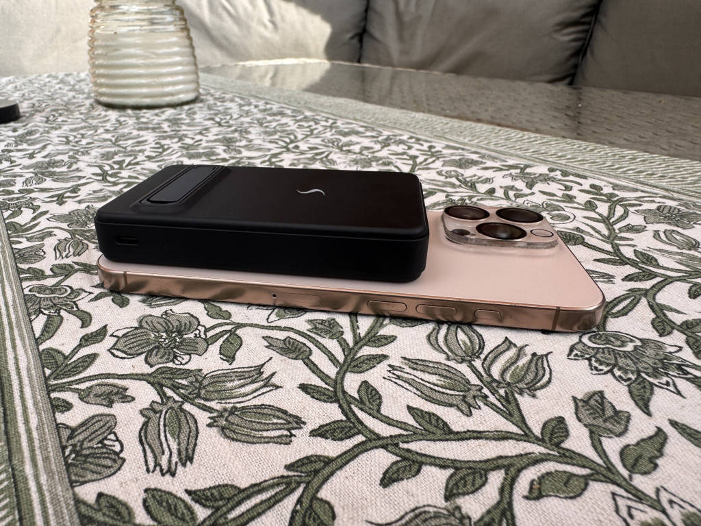 A black power bank rests atop a gold smartphone with three cameras, placed on a floral-patterned fabric. Soft sunlight creates a cosy, relaxed setting.