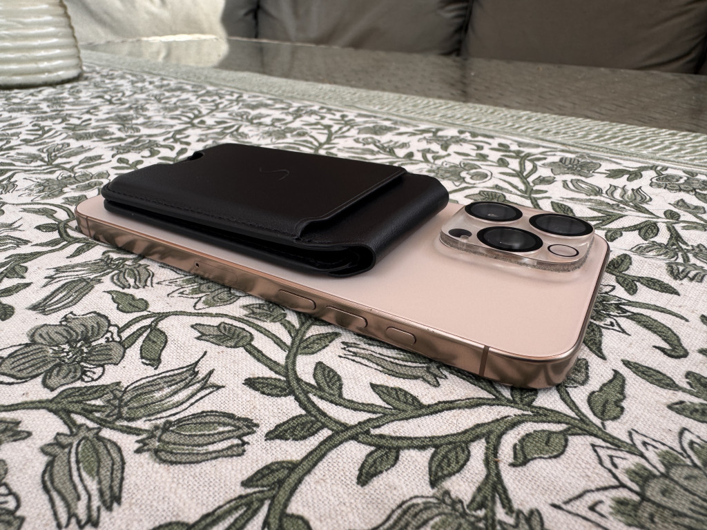 A smartphone with a black leather wallet attached rests on a floral tablecloth. The phone's camera lenses are prominent, and the setting feels casual.