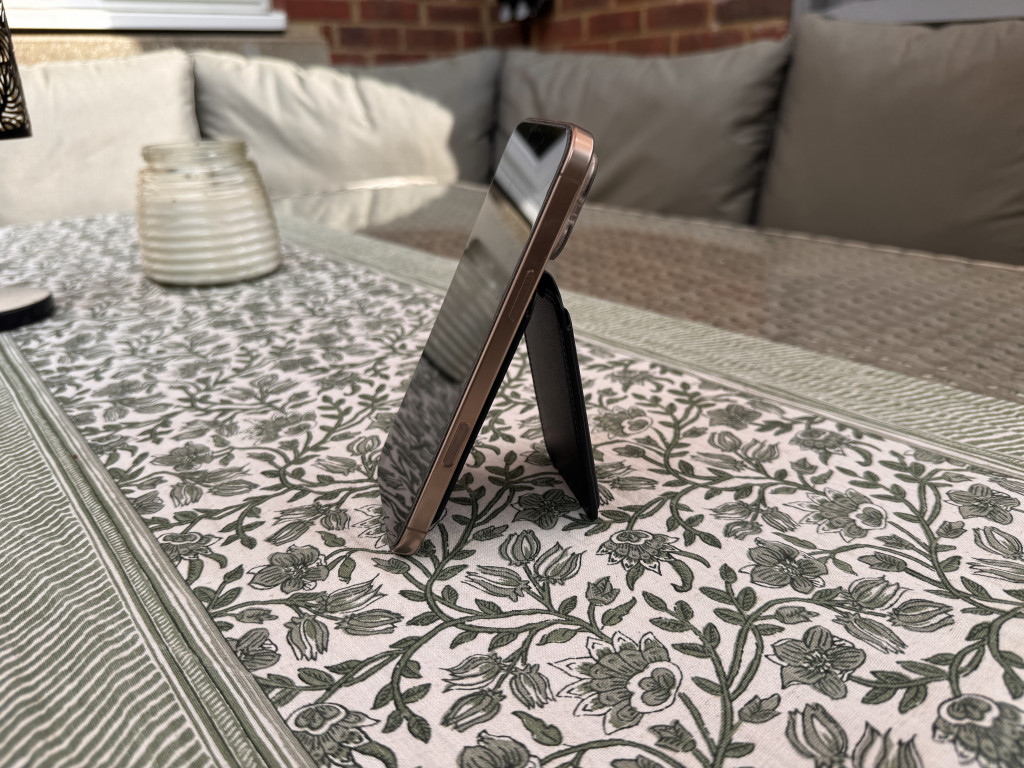 Smartphone propped up on a floral tablecloth with a stand, beside a patterned glass candle holder, on an outdoor patio with cushions. Cosy atmosphere.
