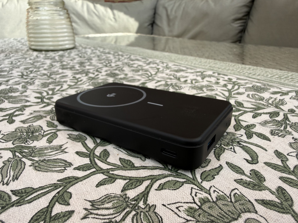 A black wireless charger sits on a floral-patterned tablecloth outdoors, with a glass jar in the background. The setting feels peaceful and relaxed.