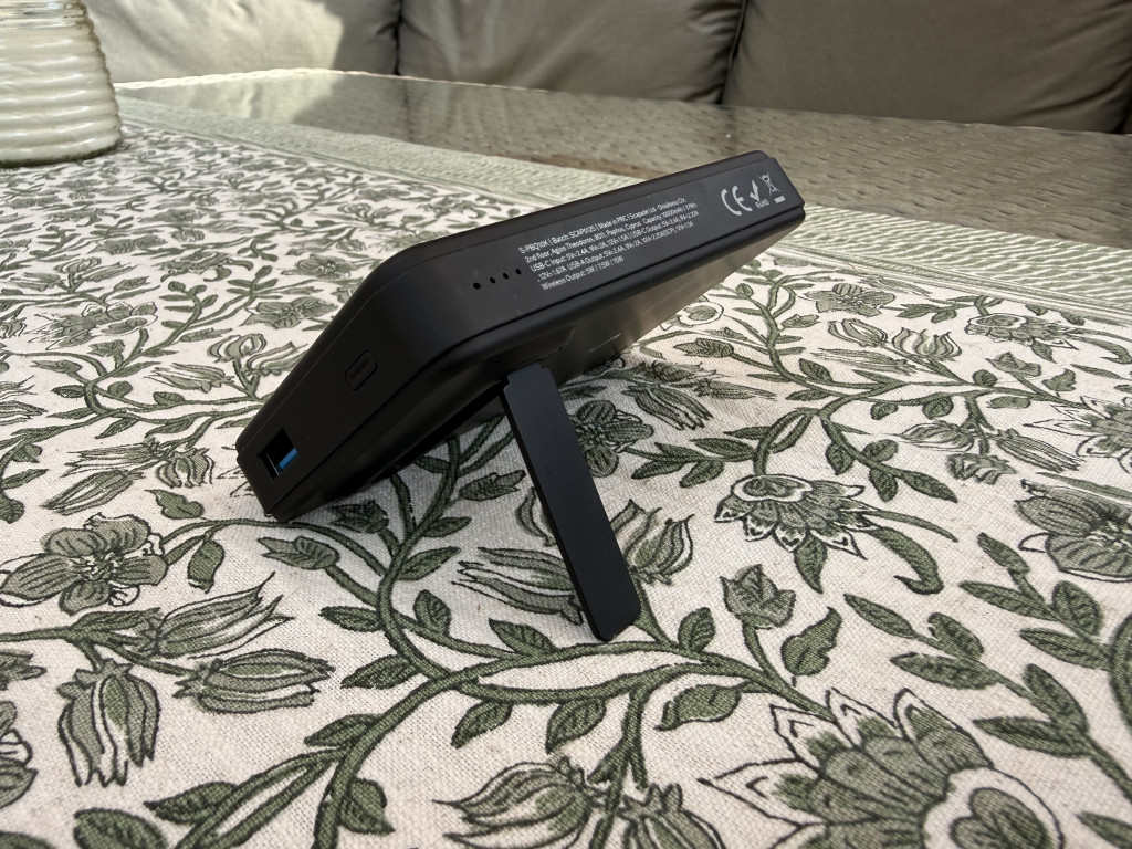 A black electronic device with a kickstand is propped on a floral-patterned tablecloth. Sunlight softly illuminates the scene, creating a relaxed ambiance.