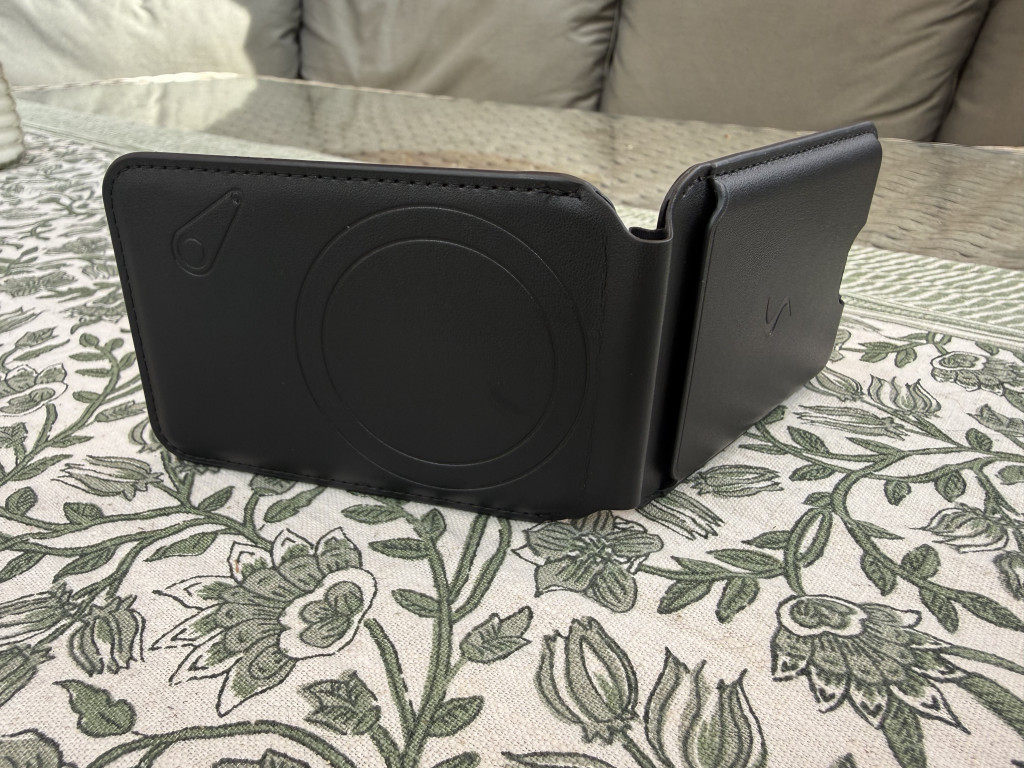 A black leather phone stand on a floral-patterned tablecloth. The stand features circular imprints and simple logo designs, conveying elegance.