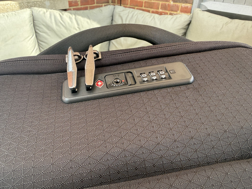Close-up of a black textured suitcase with a TSA combination lock and zip pullers. The suitcase is on a beige couch with a brick wall backdrop.