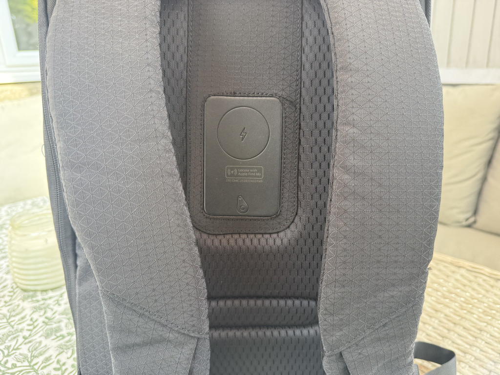 Close-up of a backpack's back panel focusing on a wireless charging symbol on a black pad nestled between grey padded straps. The setting is an outdoor patio.