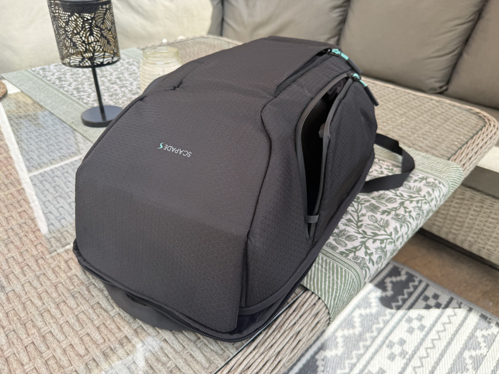 A sleek black backpack sits on a patio table, adorned with a woven pattern. It features a handle, zippers, and rests on patterned textiles, exuding a modern vibe.