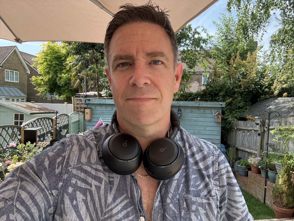 Man in a patterned shirt with headphones around his neck stands in a sunny garden. Behind him are plants, a shed, and a wooden fence, creating a serene outdoor setting.