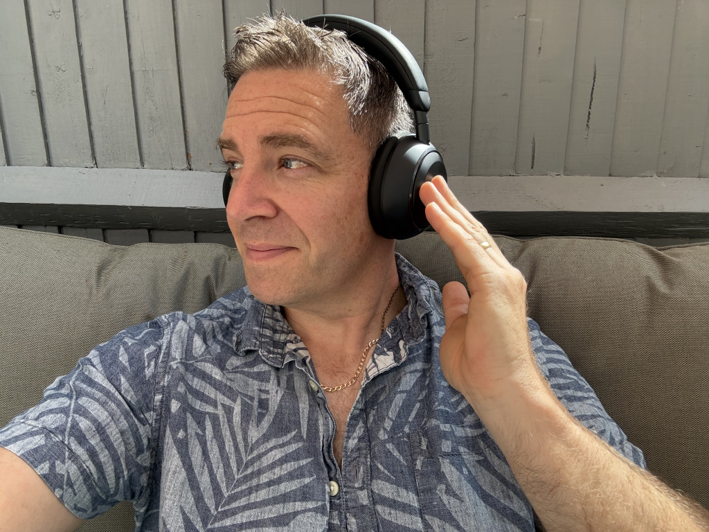 A person in a floral shirt wearing black headphones, sitting on a couch, looks contently to the side, touching the headphones, against a gray wall.
