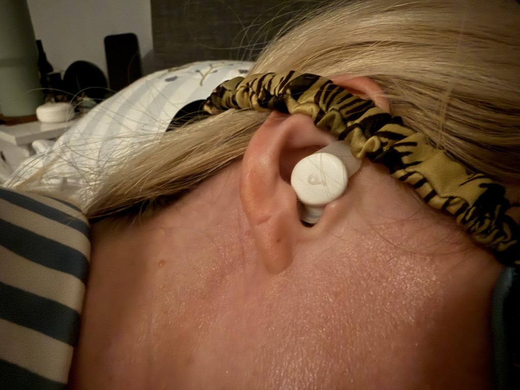 Close-up of a person lying on their side with a white earbud inserted and a patterned headband around their head. The setting appears to be cosy and relaxed.