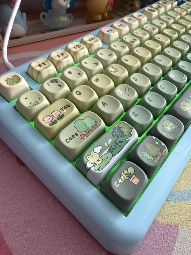 A KiiBOOM phantom 81 lite keyboard with the KiiBOOM X Cosydiaries Rainy Froggy Day keycap sets.A blue keyboard with green keycaps featuring cute frog and nature-themed illustrations with a glowing green backlight