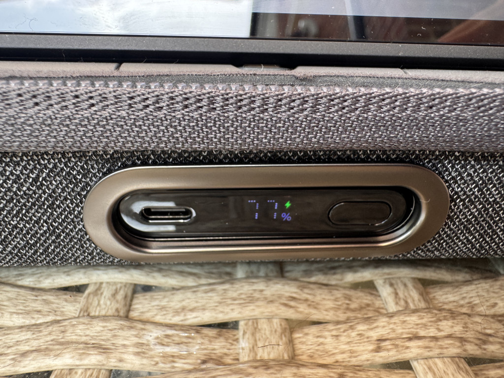 Close-up of the Belkin charging case pro with a fabric texture. A display shows battery charging at 77% near a USB-C port. Mood is tech-focused and modern.