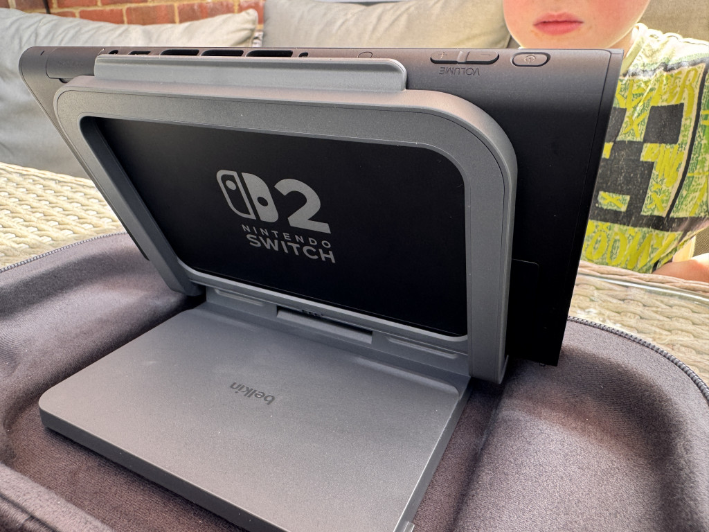 Nintendo Switch docked in a grey Belkin stand on a textured surface. A child in a yellow patterned shirt is partially visible in the background.