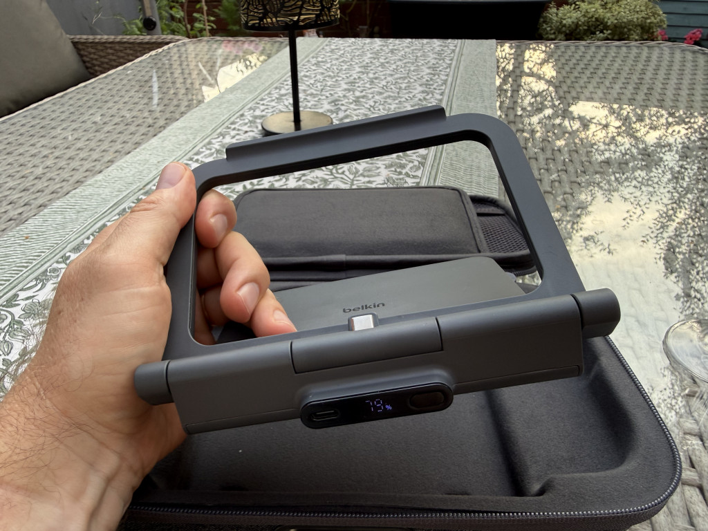 A hand holds a grey Belkin battery with a rectangular support frame, showing a digital display of the battery percentage and USB ports. Background includes a patterned outdoor table.