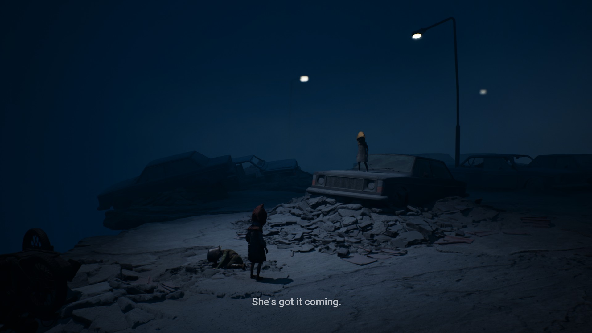 A crumbled highway in the night with many cars seen around, some are falling just a little off its edges. One car in the middle has a small child wearing a yellowish mask, while two are seen closer to the screen. A child is laying on the floor while the one next to it seems to be conversing. Some dialogue at the bottom reads "She's got it coming".