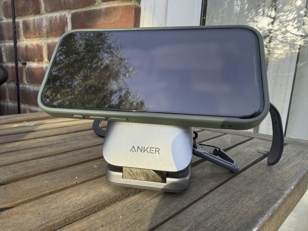 Smartphone displaying 18:46 rests on a green stand above an Anker wireless charger on a wooden table by a brick wall, outdoors.