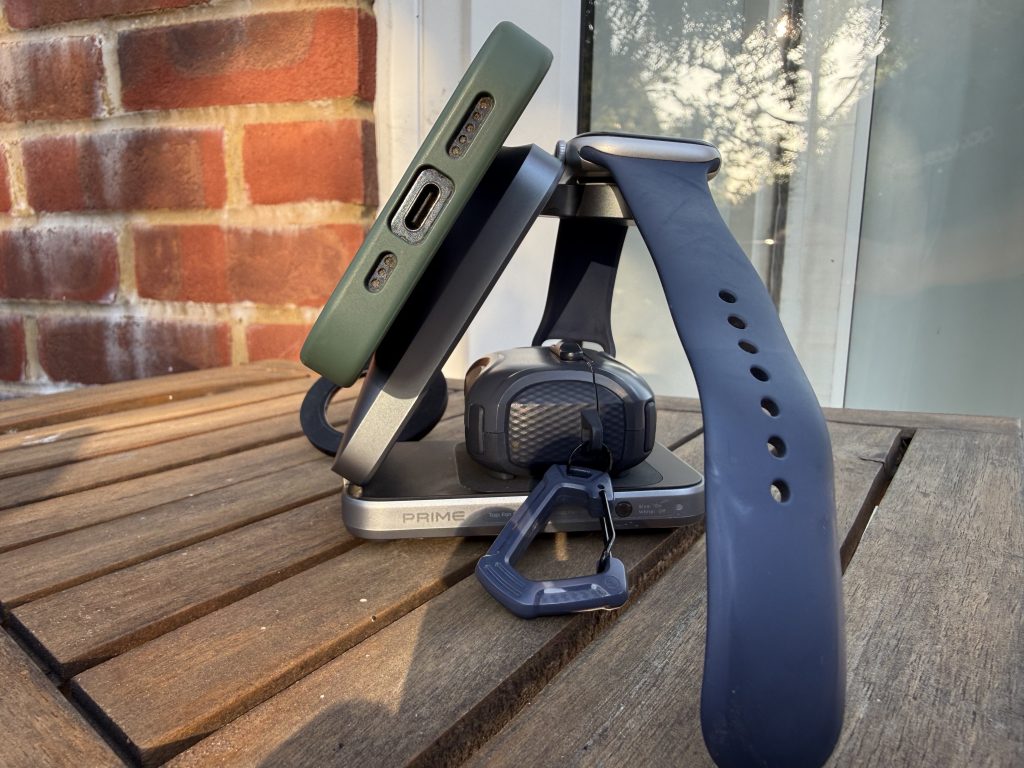 Dark blue smartwatch with perforated band rests on an Anker charging dock beside a green phone case, on a wooden table by a brick wall.