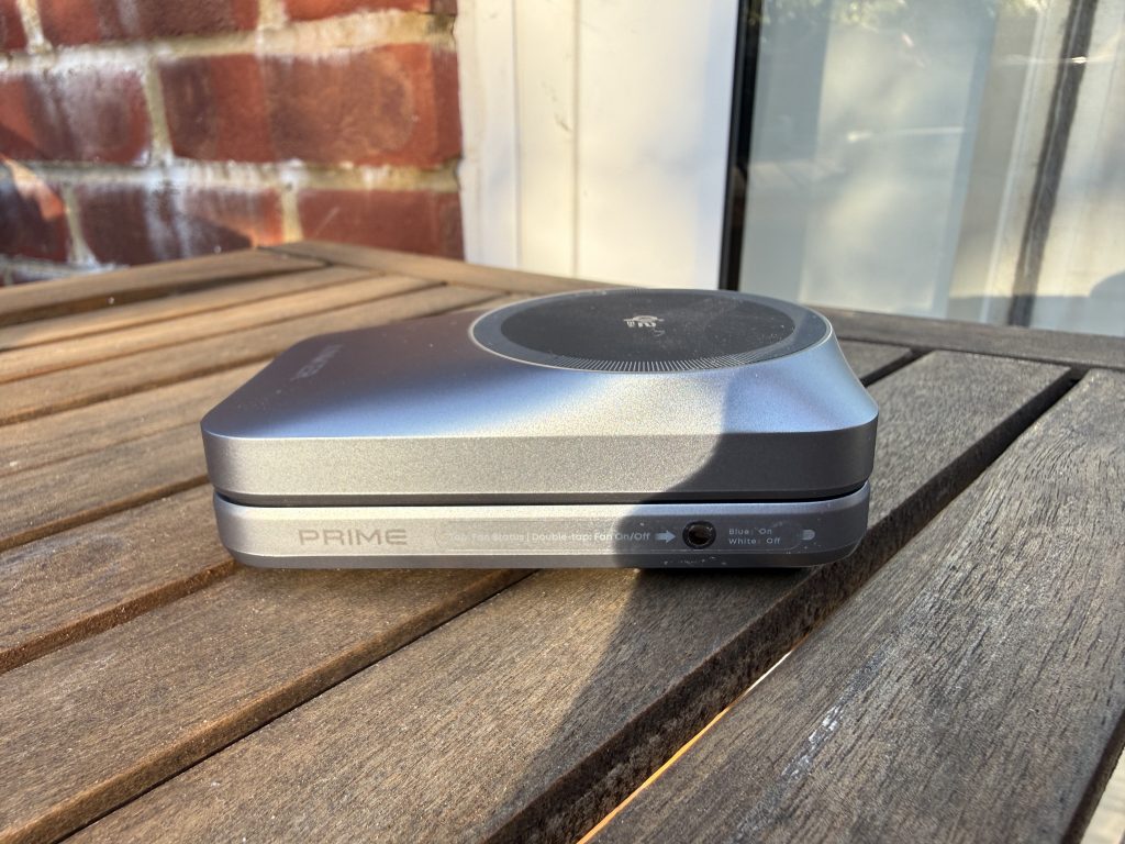Compact silver Anker “PRIME” charging device with a circular top panel sits on a weathered wooden table in warm sunlight beside a brick wall.